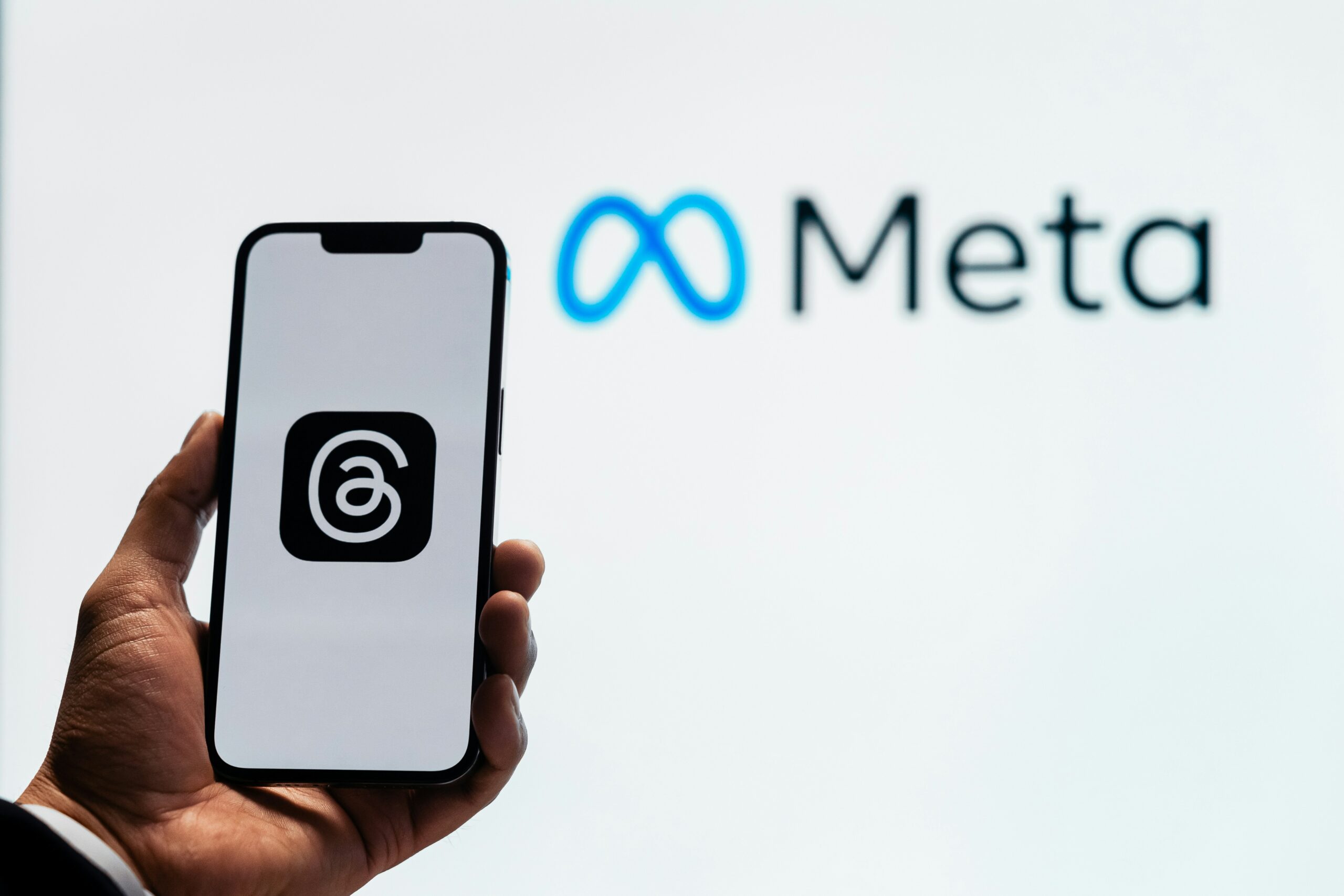 A phone holding the logo for Meta's threads in front of a white screen displaying the Meta logo